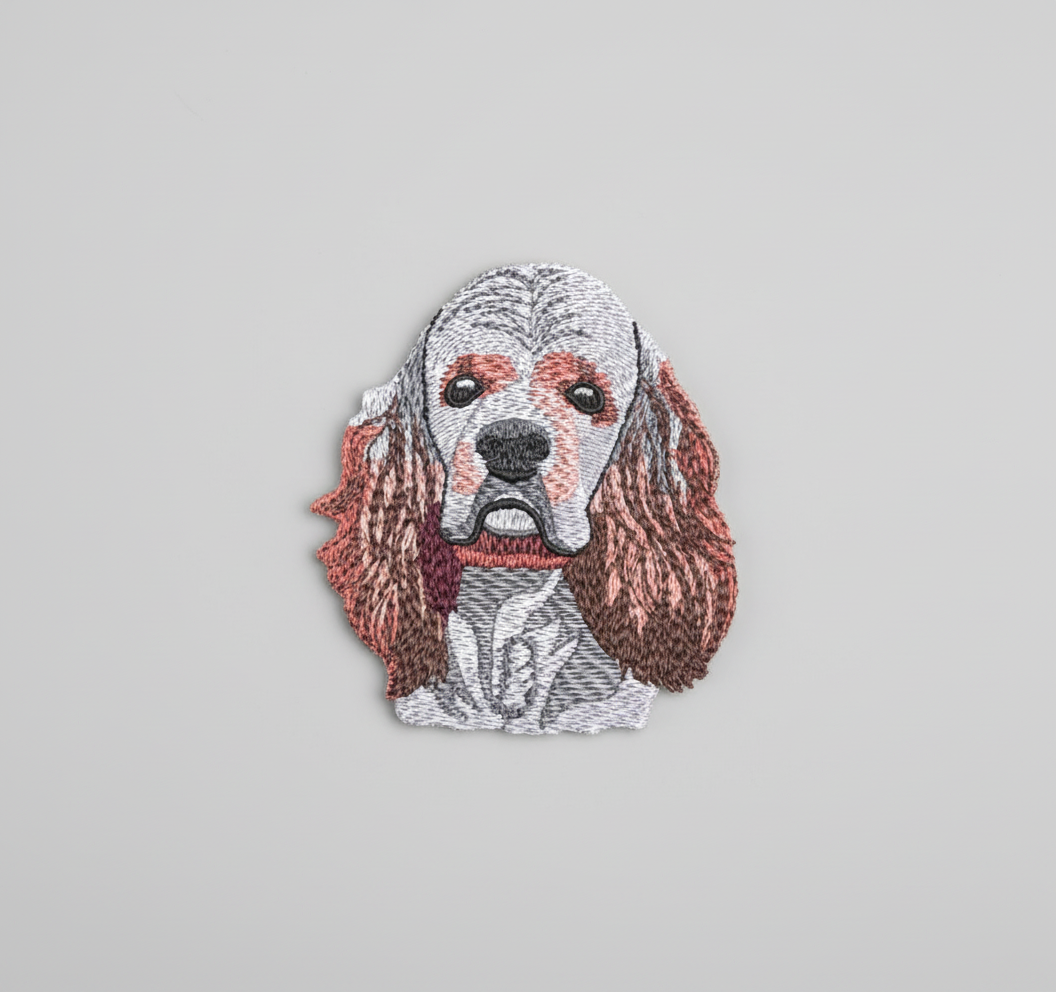Realistic American Cocker Spaniel embroidery patch featuring a detailed dog face with long floppy ears in white, brown, and rust tones, stitched with textured thread shading on a light background, original EmbroMania artwork.