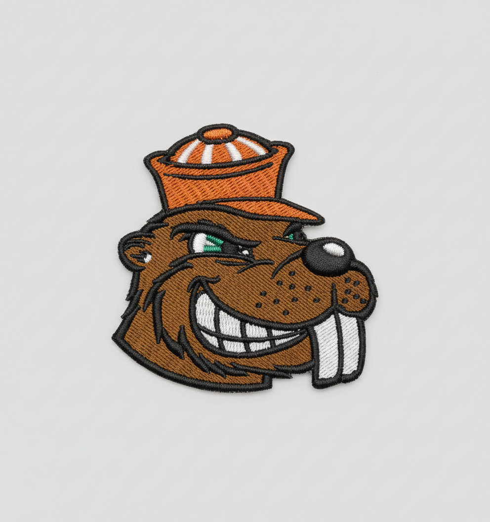 Beaver embroidery patch featuring a smiling beaver head mascot with brown fur, big white teeth, green eye detail, and an orange hat in dense stitching with a bold black border, on a light background, original EmbroMania artwork.