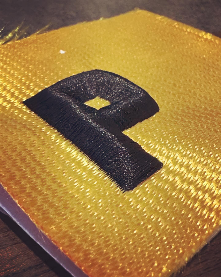 Close-up of a black 3D puff embroidered letter “P” stitched on gold fabric, showing raised texture and satin stitching.