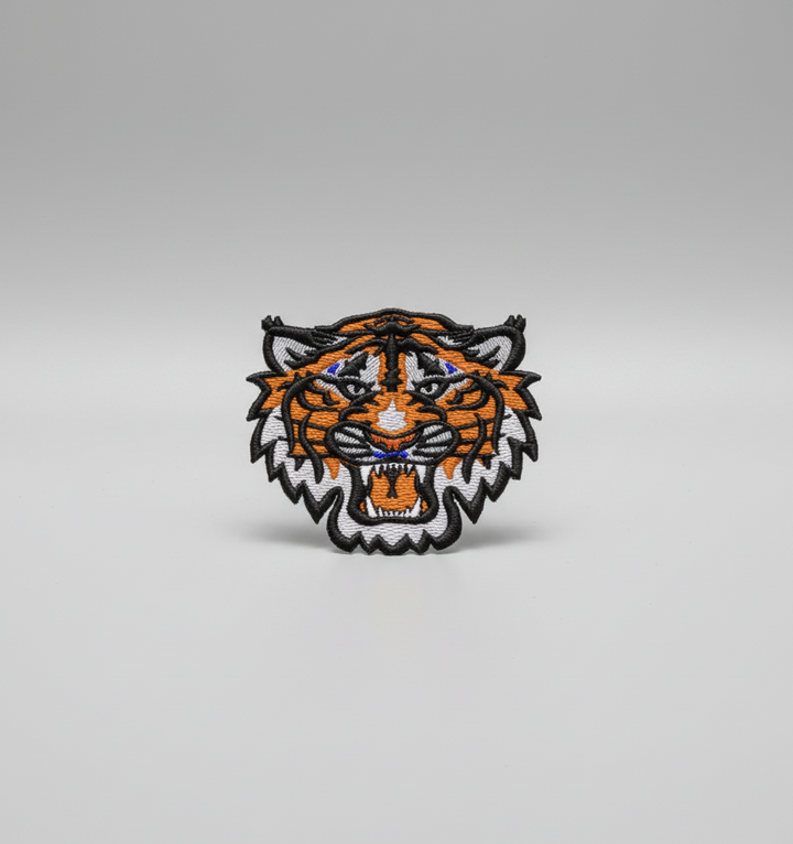 Tiger embroidery patch featuring a roaring tiger face with orange and black stripe details, white fur accents, and bold outline stitching, on a light background, original EmbroMania artwork.